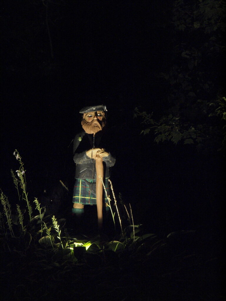 A statue of a man in a kilt standing in a wooded area at night lit up by outdoor lighting.