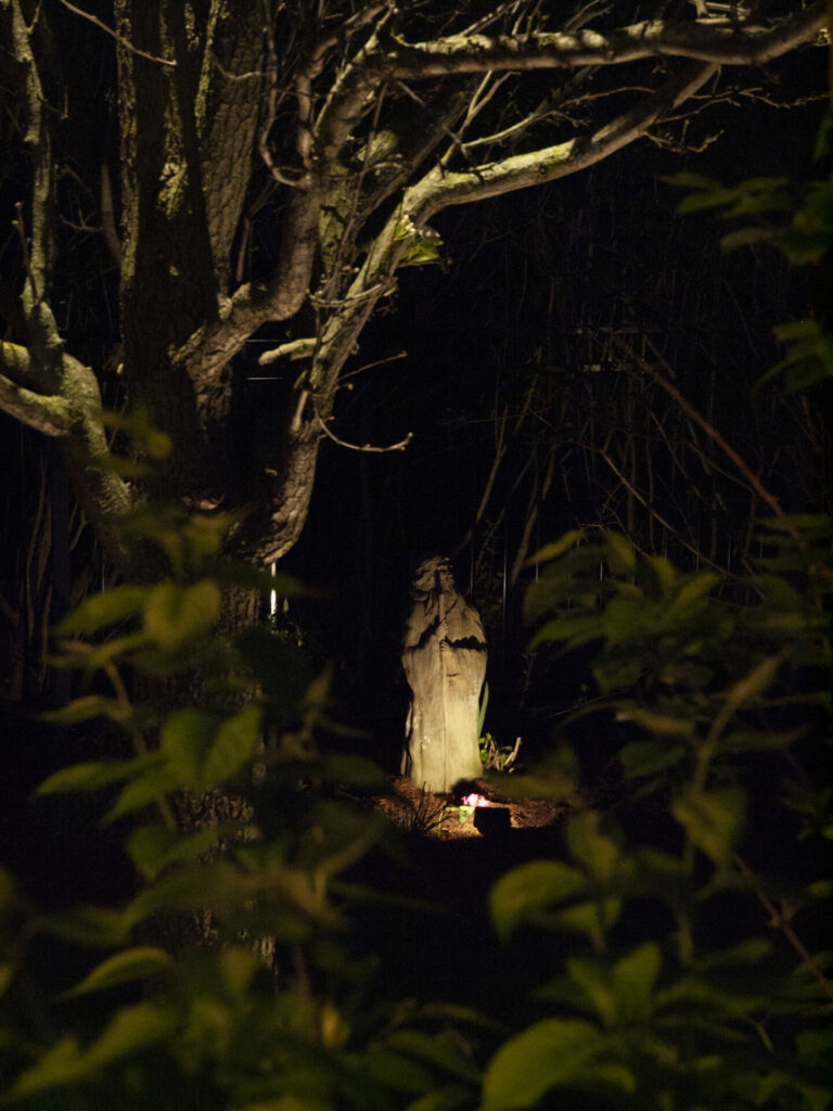 A statue in a wooded area at night lit up by outdoor lighting.