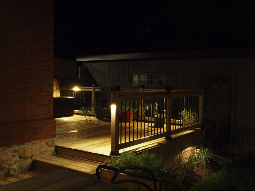 A wooden deck and stairs with potted plants lit up by outdoor lighting at night.