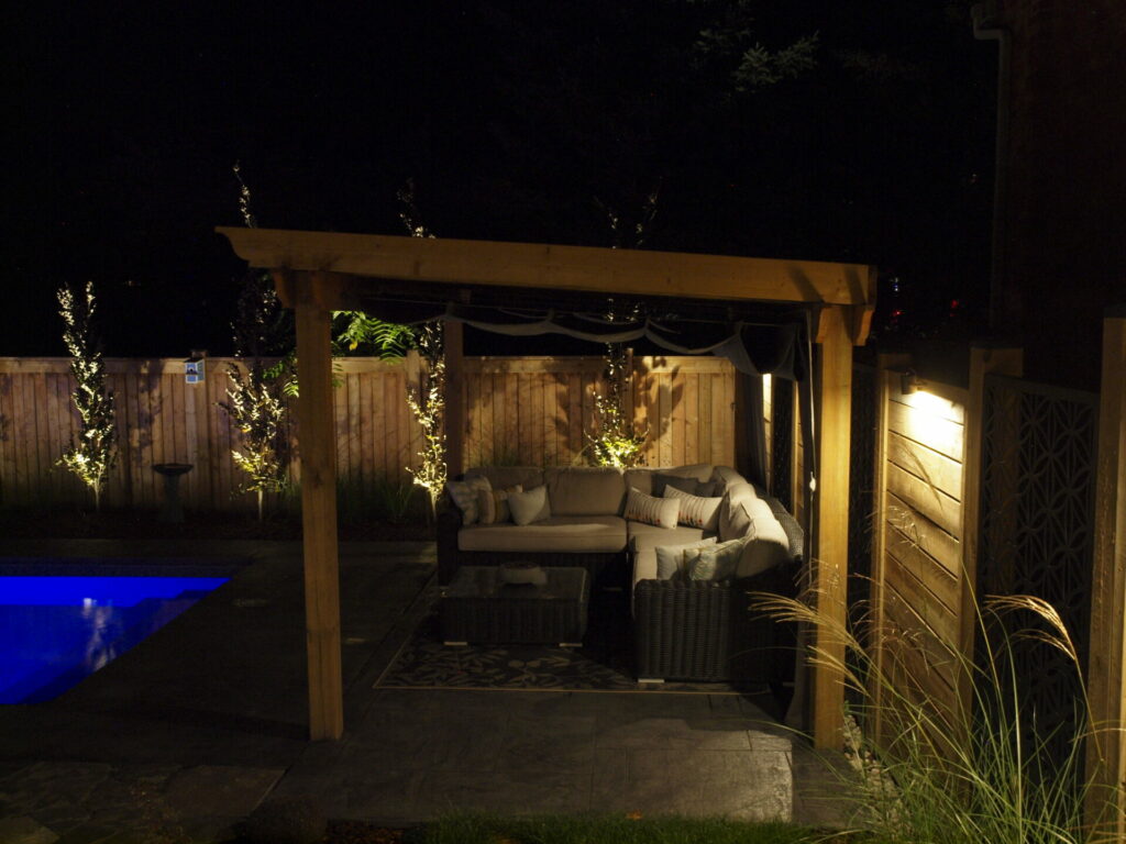 A wooden pergola next to a swimming pool lit up by outdoor lighting at night.