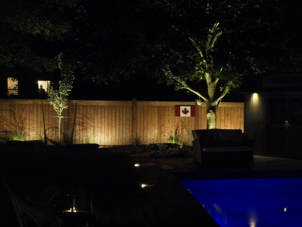 A swimming pool lit up at night with trees and a Canadian flag on a wooden fence.