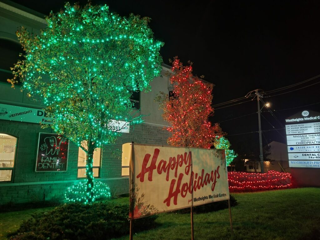 A sign that says happy holidays in front of a building and red and green lights on trees and shrubs.