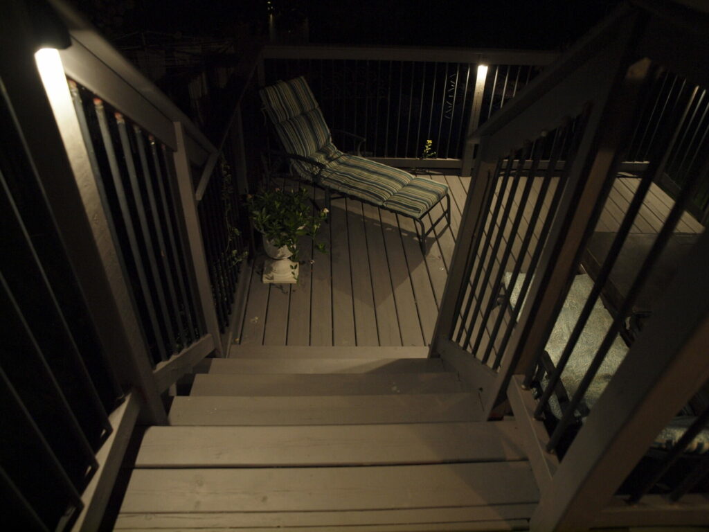 A wooden stairway with outdoor lighting in the dark leading to a deck with a lounger chair.