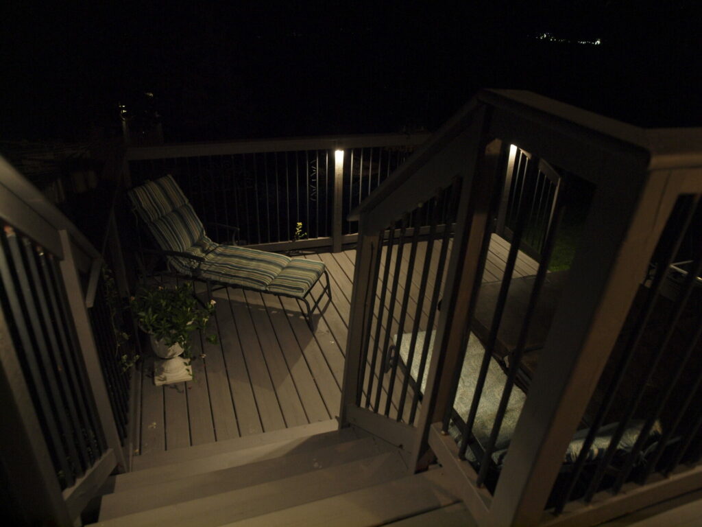 A wooden stairway with outdoor lighting in the dark leading to a deck with a lounger chair.