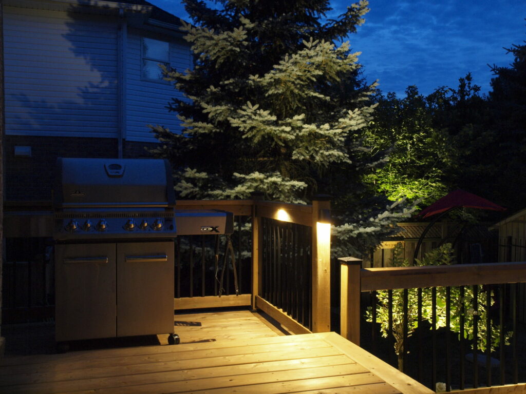 A wooden deck with a barbeque lit up at night.